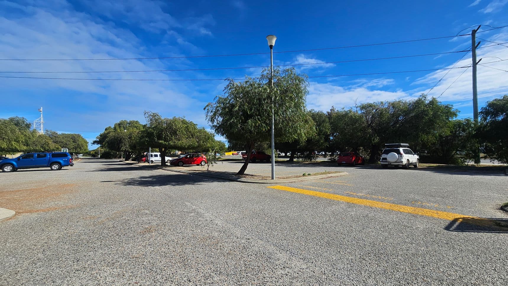 A car park with a gravel surface. On the left, a blue vehicle is parked, while on the right, a white vehicle is visible. In the centre, a tall streetlamp stands next to a tree. Several other trees and parked cars are scattered throughout the background. Power lines stretch across the top of the image, and a yellow painted line is visible on the ground in the foreground.