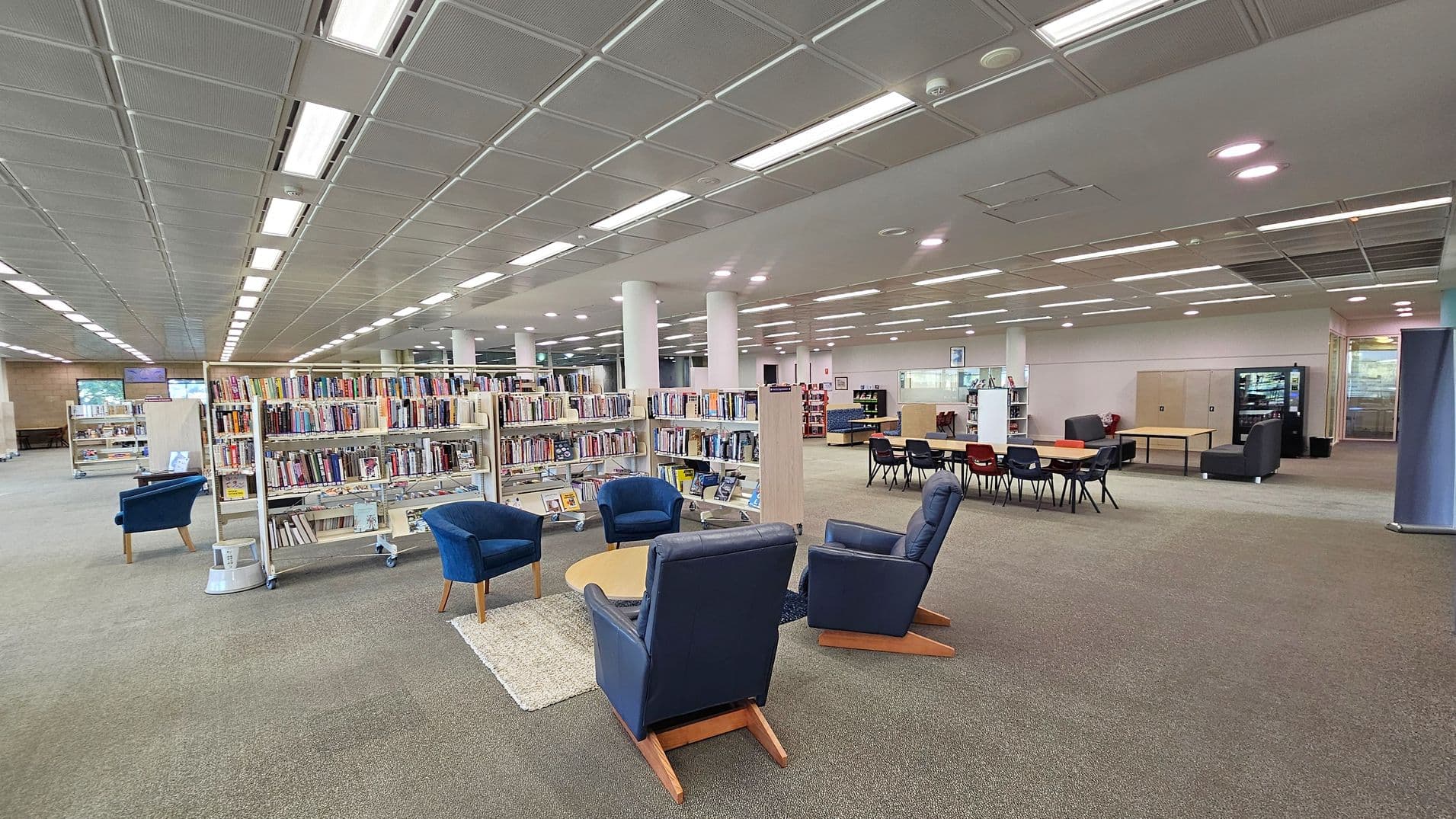 A spacious library interior with bookshelves filled with books in the centre. On the left, there are blue armchairs around a small round table on a rug. To the right, a seating area with tables and chairs, and a vending machine in the background. The floor is carpeted, and the ceiling features a grid pattern with fluorescent lighting. White columns are evenly spaced throughout the room.