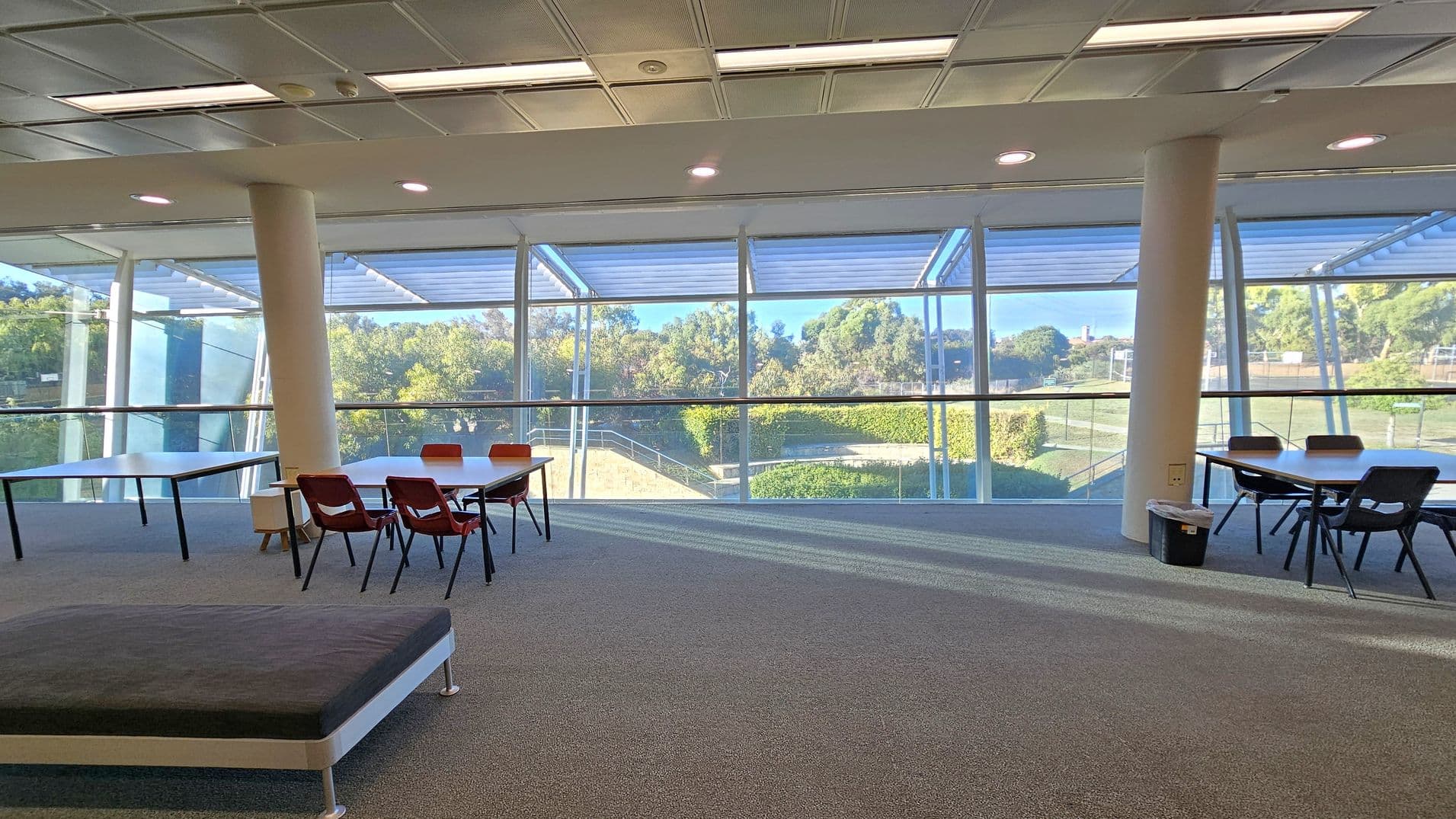 A spacious room with large floor-to-ceiling windows in the background, offering a view of greenery outside. On the left, there are tables with red chairs, and on the right, more tables with black chairs. The floor is carpeted in grey, and the ceiling has recessed lighting and a grid pattern. Two white columns are visible, supporting the structure.