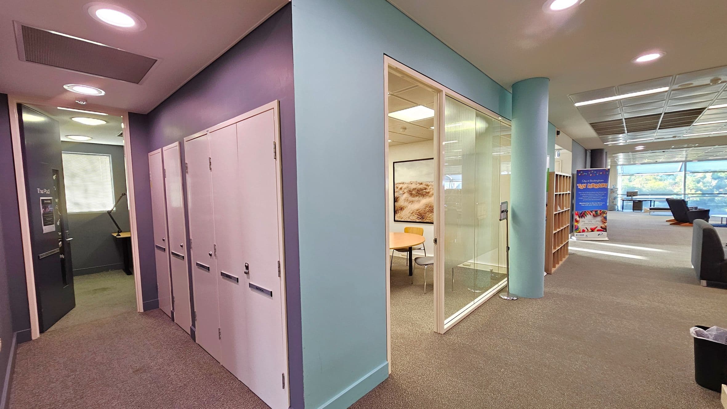 A modern office space with a corridor on the left featuring white lockers and a door labelled "The Pod" leading to a small room with a desk and lamp. In the centre, a glass-walled meeting room contains a round table and chairs. On the right, an open area with bookshelves, a colourful banner, and seating near large windows. The ceiling has recessed lighting, and the floor is carpeted.