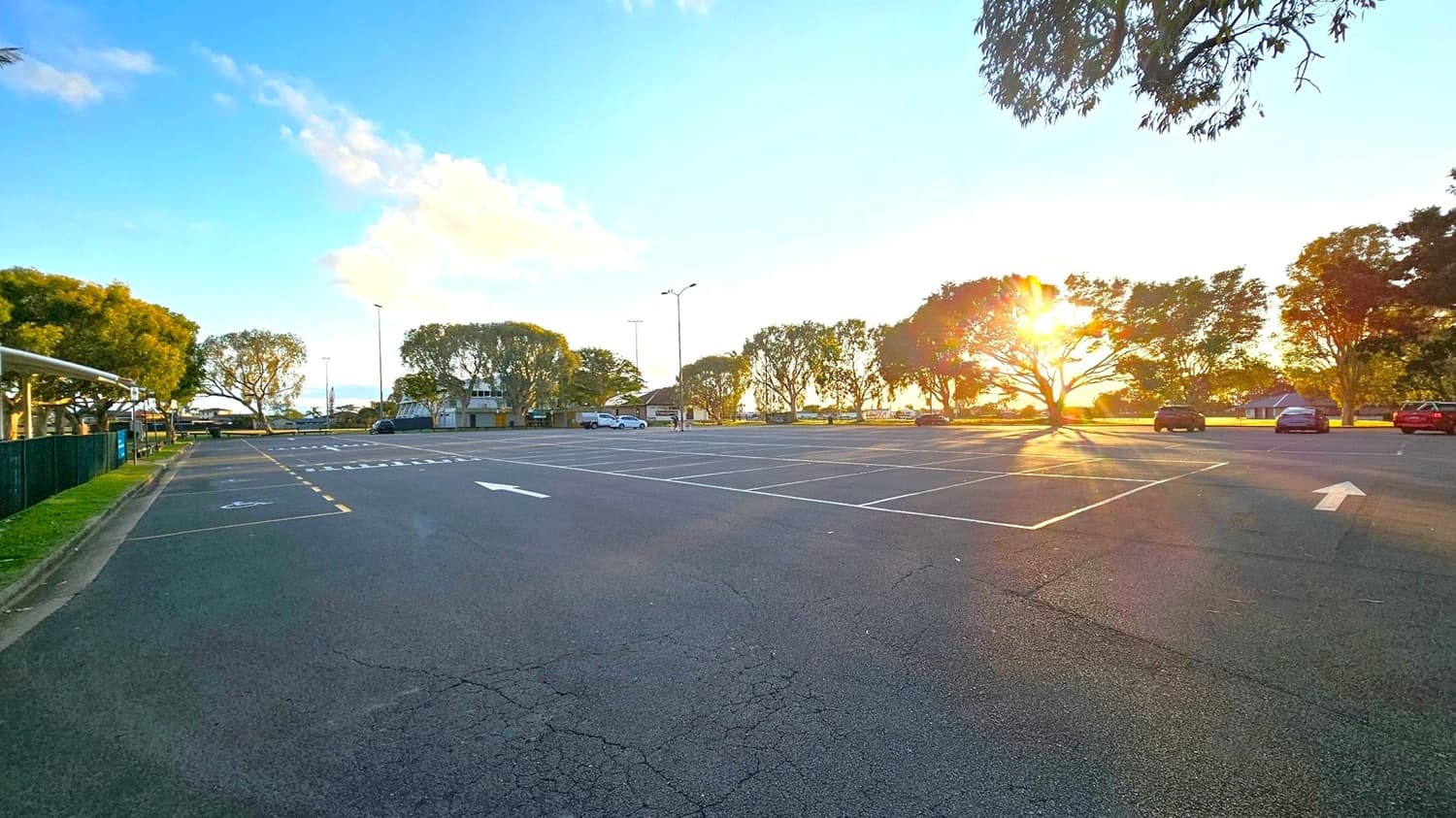An empty car park with marked spaces and a few parked cars on the right. The sun is setting behind a row of trees, casting long shadows across the asphalt. On the left, a green fence runs alongside a grassy area. In the background, more trees and a few buildings are visible. The sky is clear with some clouds, and the overall scene is calm and quiet.