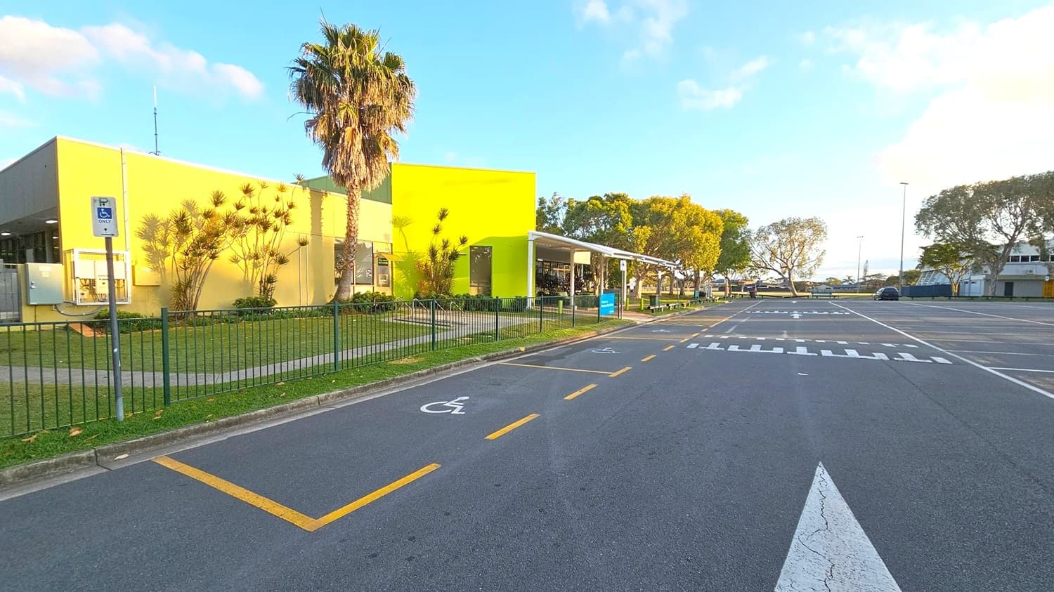 A road with marked parking spaces, including a designated disabled parking spot on the left. A bright yellow building with a palm tree and other plants in front is on the left side, enclosed by a green fence. The building has a flat roof and large windows. On the right, there are more trees and a few parked cars. The road has a zebra crossing and directional arrows. The area is well-lit, suggesting daytime.