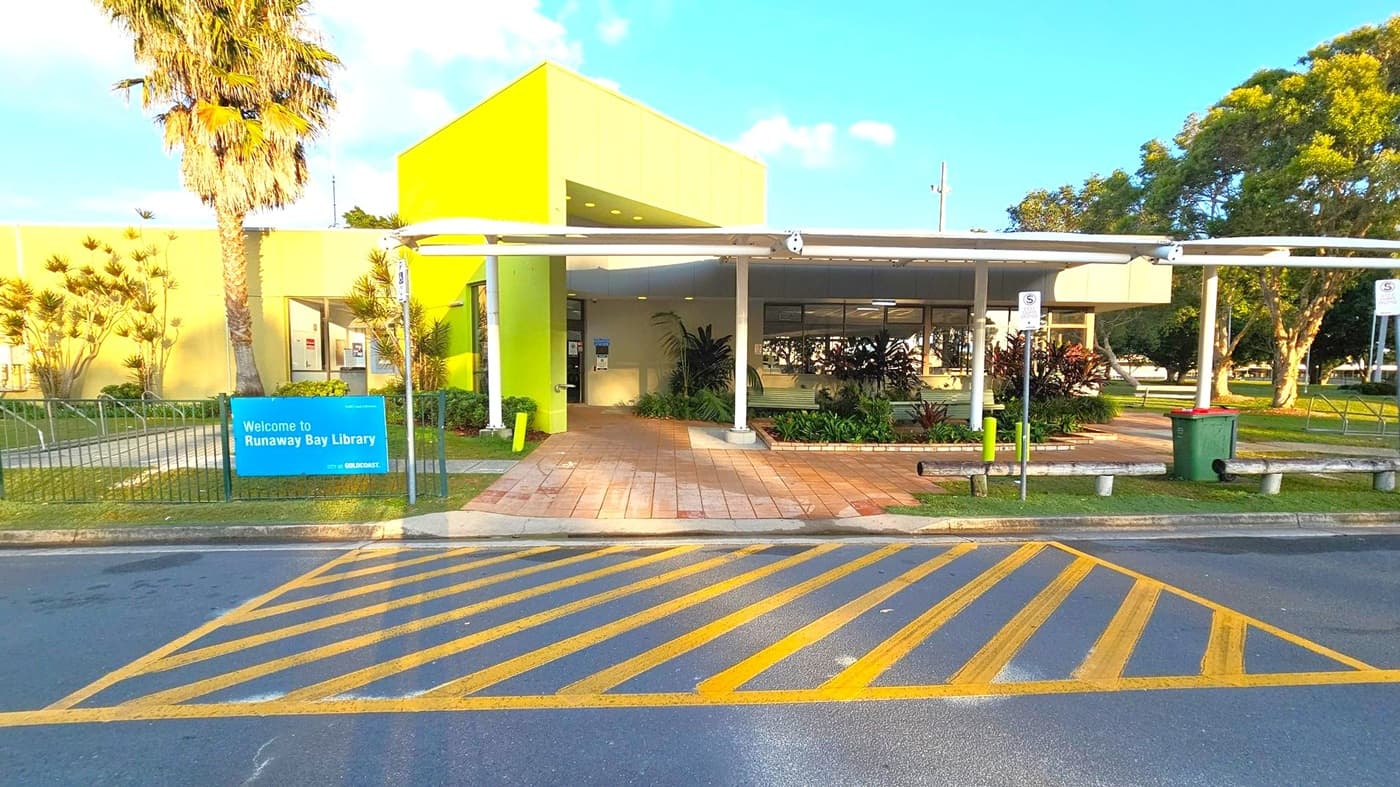 A library entrance with a bright yellow-green facade. A blue sign on the left reads "Welcome to Runaway Bay Library." A palm tree and other plants are near the entrance. The path leading to the entrance is paved with tiles. A covered walkway extends from the building, supported by white poles. Yellow diagonal lines are painted on the road in the foreground. A green bin and parking signs are on the right, with trees and grass in the background.