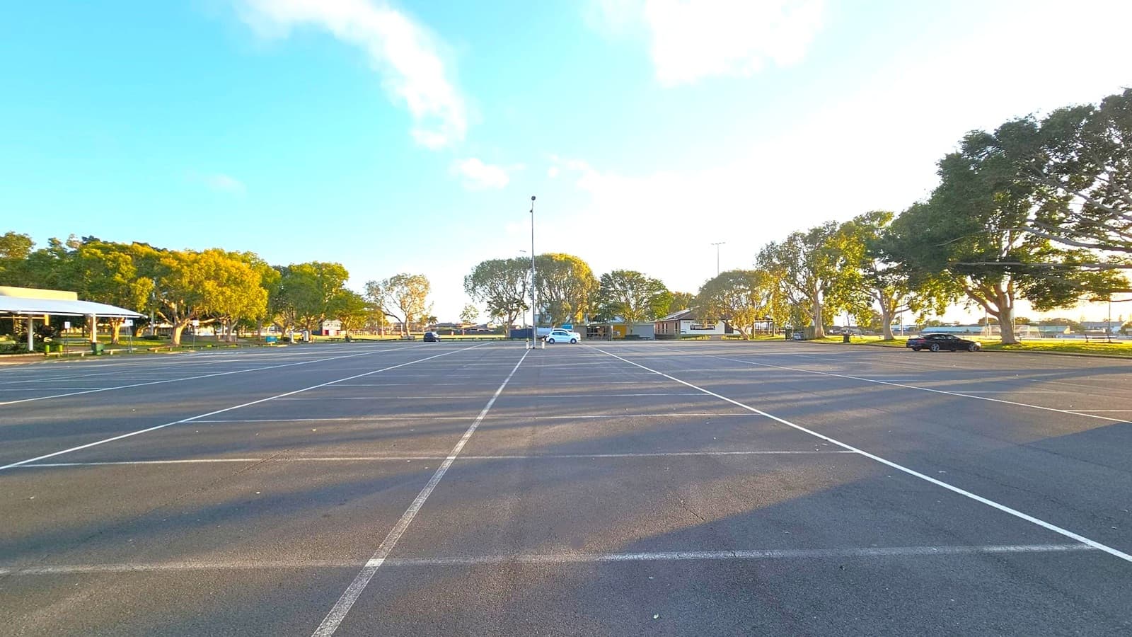 An expansive car park with marked spaces, mostly empty except for a few cars scattered around. Trees line the background, providing shade and greenery. On the left, a building with a white roof is partially visible. The ground is paved with asphalt, and the area is well-lit by natural light. A tall light pole stands in the centre, and the scene is calm and open.