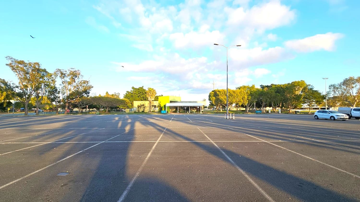 An empty car park with marked spaces stretches across the foreground. In the centre, a building with a bright green facade is visible, surrounded by trees. To the right, a few parked cars are seen. Tall streetlights cast long shadows across the ground. Trees line the left and right sides, with a few birds flying above. The sky is clear with some clouds.
