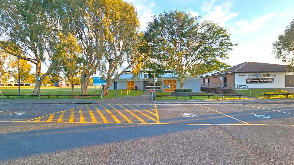 A building with a sign reading "Runaway Bay Soccer Club" on the right. In the centre, a pathway leads to the entrance, flanked by trees. On the left, a grassy field is visible with more trees and a sign. The foreground shows a car park with yellow-striped markings and designated parking spaces.