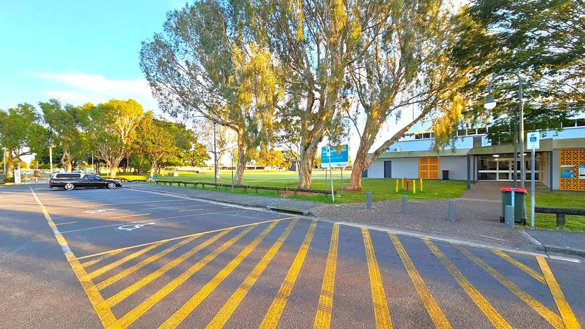 A car park with yellow diagonal lines on the ground. On the left, a black car is parked in a designated space. In the background, there are large trees with green foliage and a grassy area. On the right, a building with a grey facade and yellow decorative panels is visible. A pathway leads to the entrance, and a green bin with a red lid is nearby. A signpost and a streetlamp are also present.
