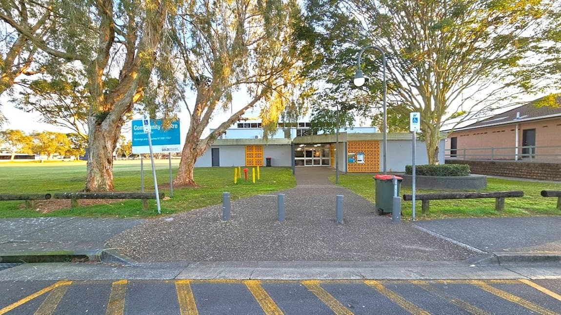 A community centre building is in the centre, with a pathway leading to its entrance. On the left, there are large trees and a sign indicating the centre's name. On the right, a lamp post and a circular planter with a tree are visible. A bin is placed near the pathway. The foreground features a marked parking area with yellow lines, and the ground is paved. The building has a flat roof and a facade with decorative brickwork.