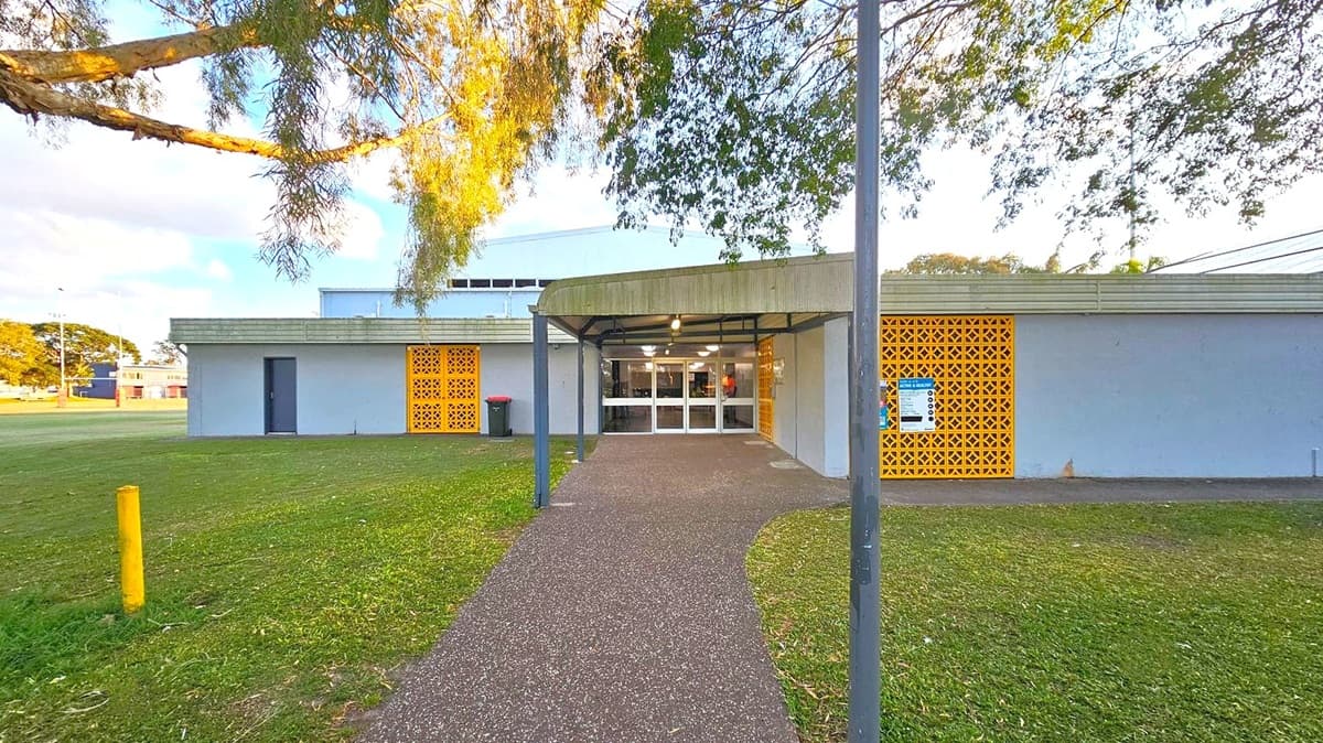 A building with a flat roof and a covered entrance in the centre. The facade features grey walls with decorative yellow lattice panels on either side of the entrance. A pathway leads to the entrance, flanked by grass on both sides. A tree branch extends from the left, casting shadows on the building. A yellow bollard is visible on the left side of the grass. The entrance has glass doors, and there are lights under the canopy.