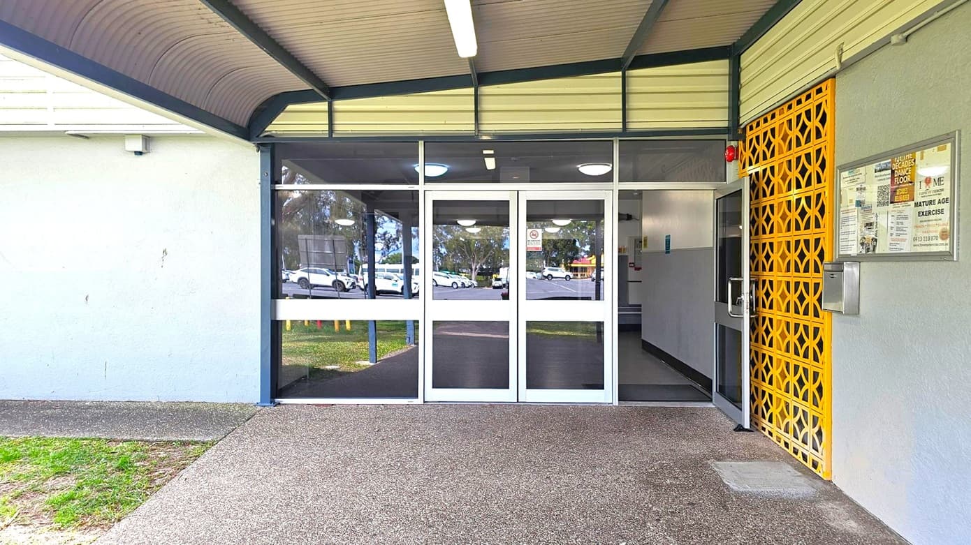 A building entrance with glass double doors in the centre, reflecting parked cars and trees outside. To the right, a yellow lattice panel and a noticeboard with various posters. The left side features a plain grey wall. The floor is a mix of concrete and grass, and the ceiling is covered with a corrugated metal awning. A fluorescent light fixture is mounted on the ceiling.