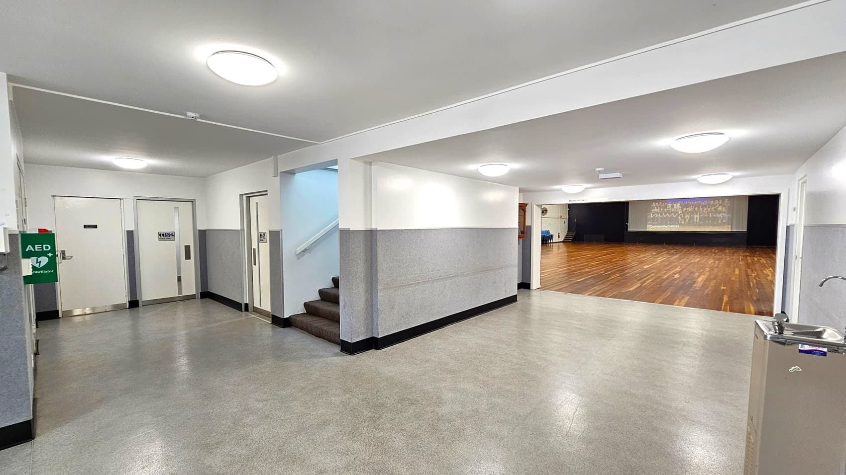 A hallway with grey flooring and white walls. On the left, there are two white doors with signs and an AED mounted on the wall. A staircase with dark steps is visible next to the doors. On the right, a water fountain is attached to the wall. The hallway opens into a large room with a wooden floor and a stage at the far end. The ceiling is white with circular lights.