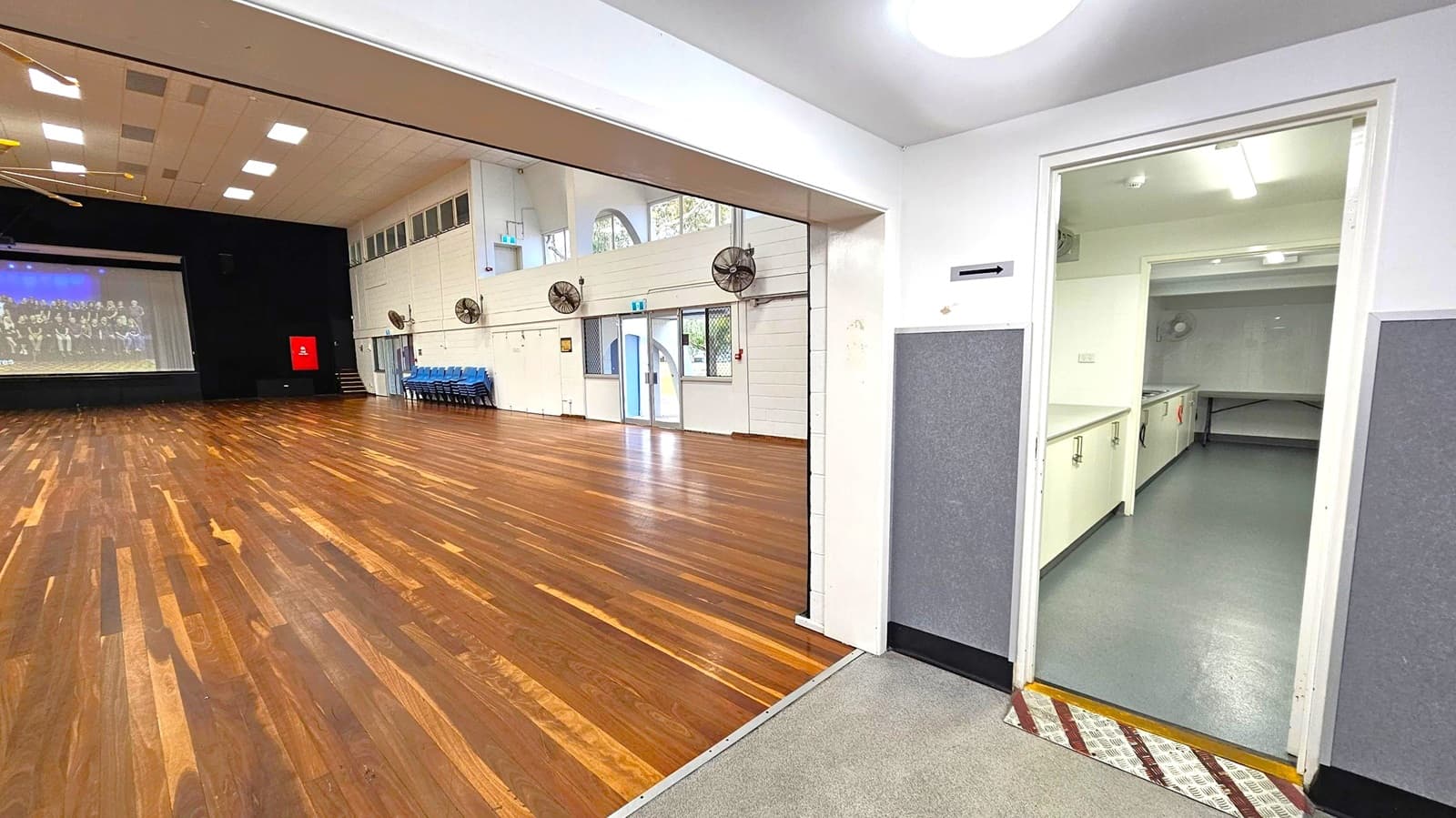 A large hall with a wooden floor and a high ceiling with square lights. On the left, a stage with a screen displaying an image. Several blue chairs are stacked against the wall. The right side shows an open door leading to a kitchen area with grey flooring and white cabinets. The walls are white with grey panels, and there are several wall-mounted fans. A door with glass panels is visible in the background, leading outside.