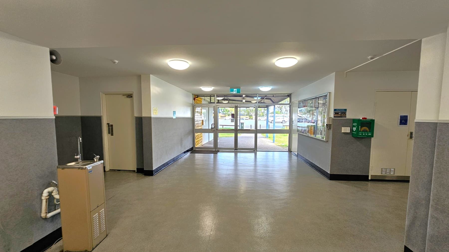 A hallway with a water fountain on the left and a door next to it. The floor is a speckled grey and green, and the ceiling is light green with round ceiling lights. On the right, there's a wall-mounted AED and a bulletin board. At the end of the hallway, glass doors lead outside, showing a view of a grassy area and parked cars.