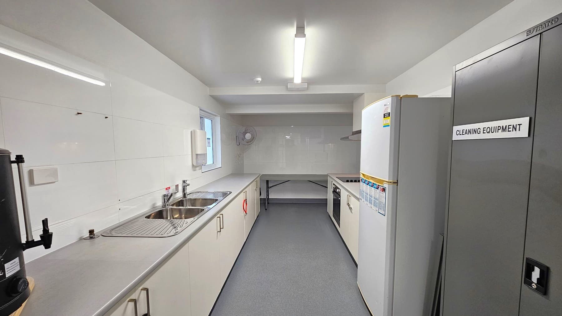 A narrow kitchen with white walls and a grey floor. On the left, there's a long countertop with a double sink and a hot water dispenser. A window is above the sink. On the right, a tall cabinet labelled "Cleaning Equipment" and a white refrigerator. Further back, an oven and stovetop are built into the counter. A small fan is mounted on the wall in the background. The ceiling has a fluorescent light fixture.