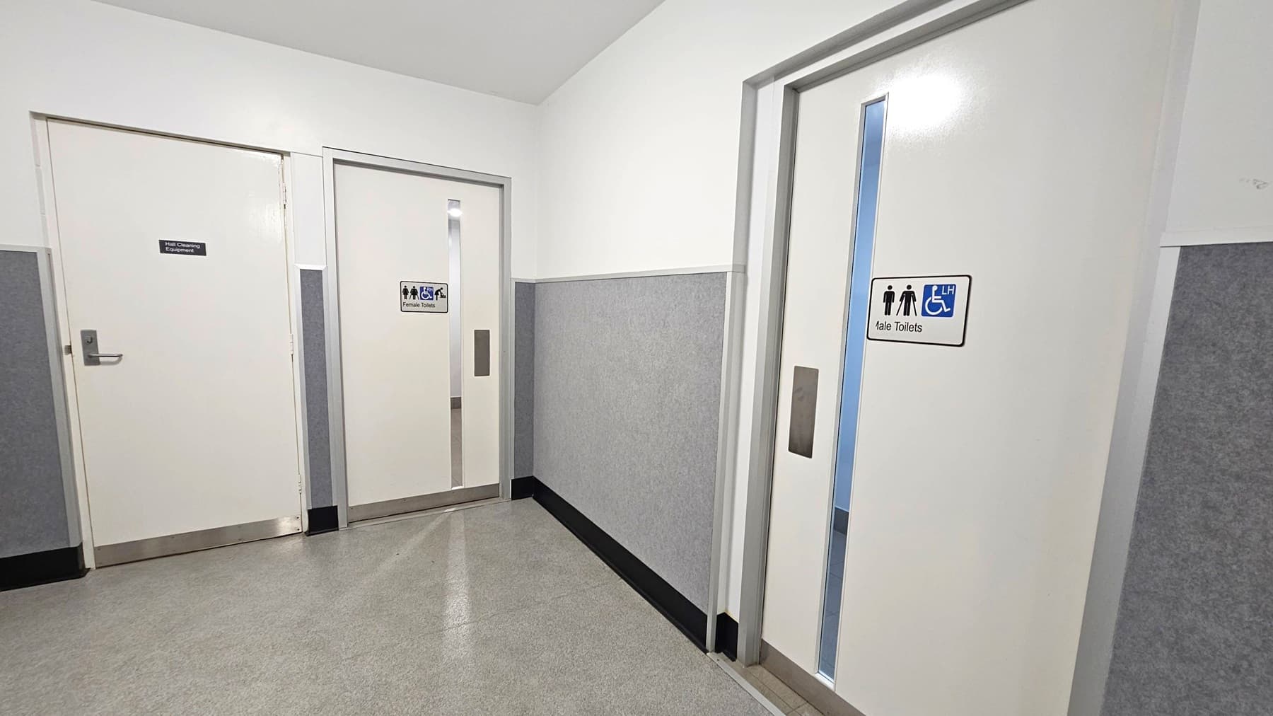A corridor with three white doors. On the left, a door with a sign for "Male Cleaners Equipment." In the middle, a door marked "Female Toilets" with symbols for accessibility. On the right, a door labelled "Male Toilets" also featuring accessibility symbols. The walls are grey with a white upper section, and the floor is covered in grey tiles. The ceiling is plain white.