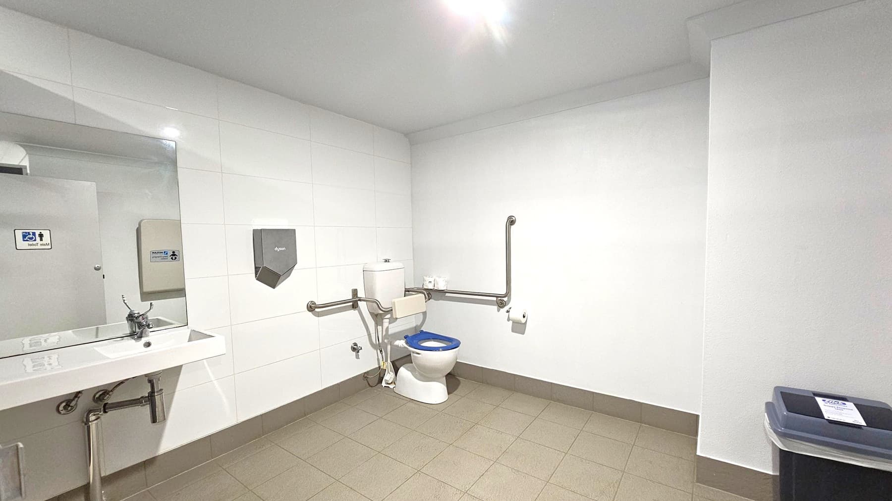 A public accessible toilet with white tiled walls and a beige tiled floor. On the left, a sink with a mirror above it and a hand dryer mounted on the wall. In the centre, a toilet with a blue seat, equipped with support rails on both sides. Toilet paper is mounted on the right rail. On the right, a bin with a lid. The ceiling is plain white with a light fixture.