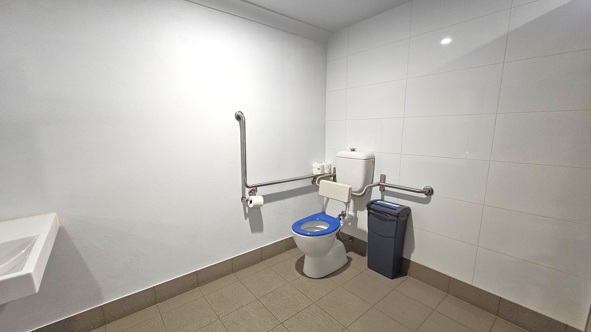 A bathroom with white tiled walls and a beige tiled floor. On the right, there is a toilet with a blue seat and a backrest, equipped with metal grab bars on both sides. Above the toilet, a cistern is visible, with toilet paper rolls placed on top. A black waste bin is positioned next to the toilet. On the left, a small white sink is partially visible. The ceiling is plain white with a recessed light.