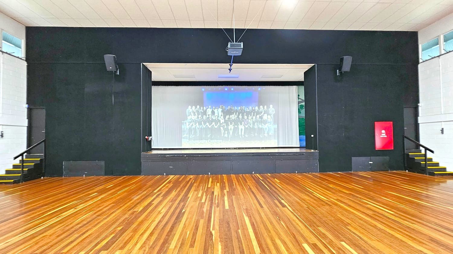 A large room with a wooden floor and a high ceiling. At the centre is a stage with a black backdrop and a projector screen displaying an image of a group of people with the text "Community Centres". There are speakers mounted on either side of the stage. On the right, a red fire safety sign is visible. Stairs with yellow-edged steps are on both sides of the stage, leading to doors. The walls are light-coloured with small windows near the ceiling.