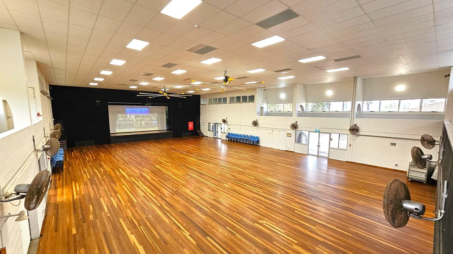 A large hall with a polished wooden floor and a high ceiling with recessed lighting. On the left, wall-mounted fans and stacked chairs are visible. The right side has more fans and a set of double doors with windows above. At the far end, a stage with a large screen displaying an image. Ceiling fans are evenly spaced across the room. The walls are light-coloured with high windows allowing natural light.