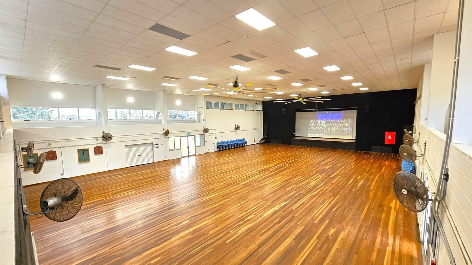 A large hall with a polished wooden floor and a high ceiling with multiple rectangular lights. On the right, several wall-mounted fans are visible. At the far end, a stage with a large screen displaying an image. Blue chairs are stacked against the wall near the stage. The left side features windows and a set of double doors. Ceiling fans are centrally located.