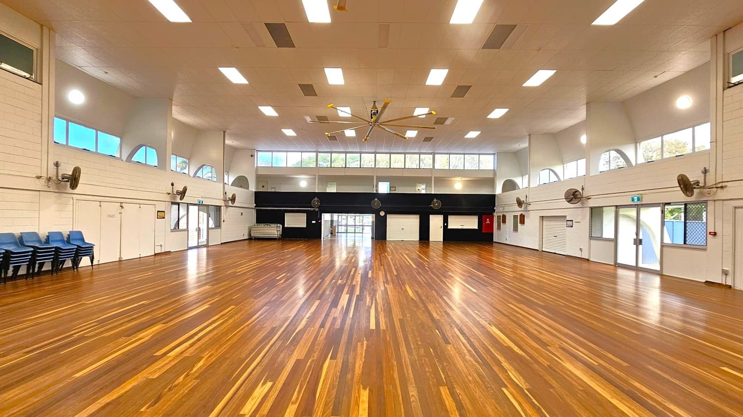 A spacious hall with polished wooden flooring and a high ceiling featuring multiple rectangular lights. The left side has stacked blue chairs and several wall-mounted fans. The right side has large windows and doors with metal grilles. The back wall has a series of doors and windows above, with a large ceiling fan in the centre. The walls are white with some black sections, and there are emergency exit signs above the doors.