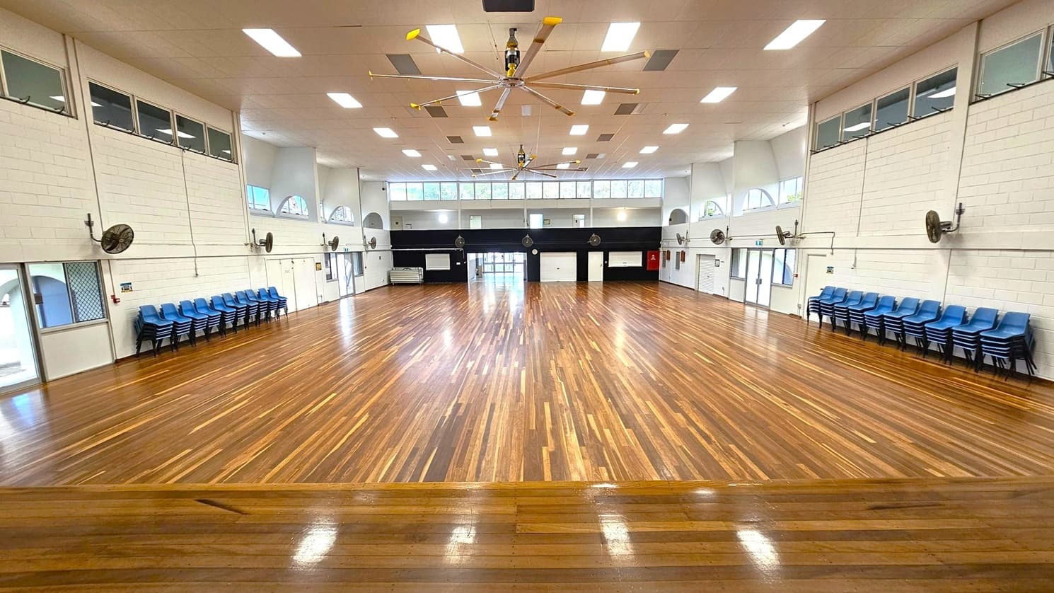A large hall with polished wooden flooring and a high ceiling featuring multiple rectangular lights. In the centre, a large ceiling fan is visible. The walls are white with high windows and several wall-mounted fans. Stacks of blue chairs are lined up against the left and right walls. At the far end, there are doors and windows, with a black section of wall above them.
