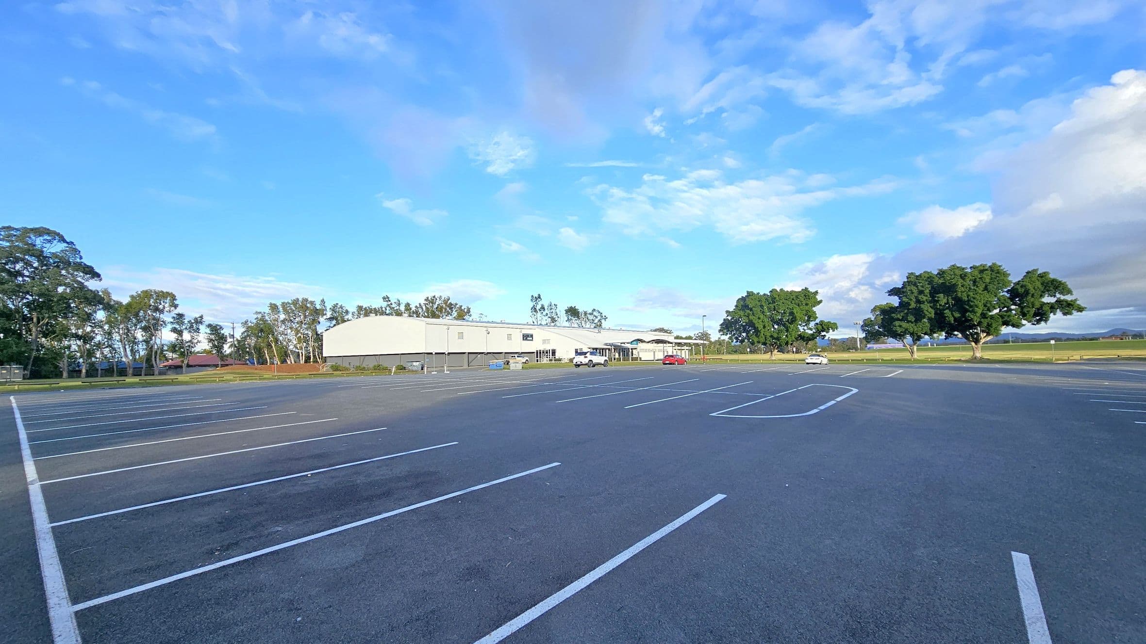 An expansive, empty car park with marked parking spaces on an asphalt surface. In the background, a large, light-coloured building is visible, with a few vehicles parked near it. To the right, there are several large trees and open grassy areas. The left side features more trees and a glimpse of another structure. The sky is clear with some clouds.