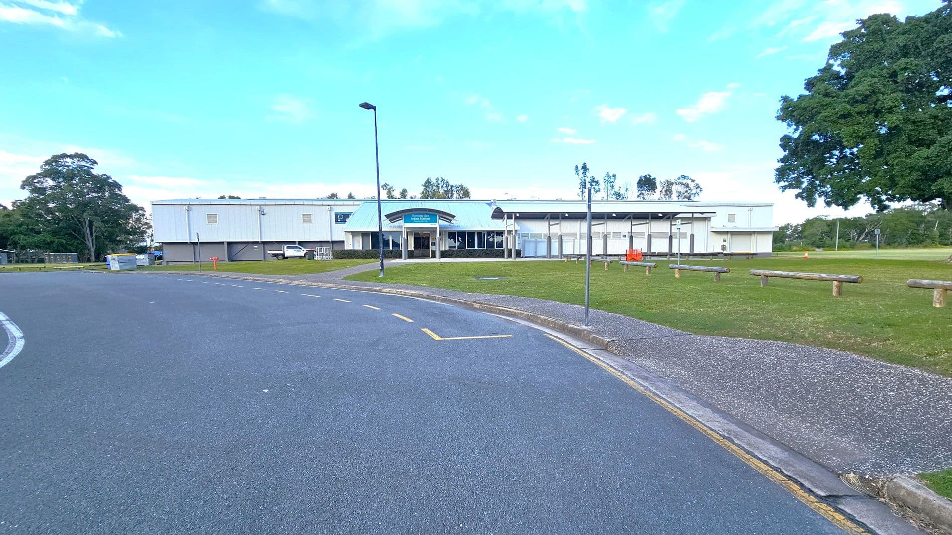 A large, single-storey building with a light-coloured exterior and a central entrance is in the background. The foreground features a wide asphalt road with yellow markings, curving slightly to the left. On the right, there is a grassy area with wooden benches and a few trees, including a large tree on the far right. A lamppost and a few signs are near the building, and a white vehicle is parked to the left.