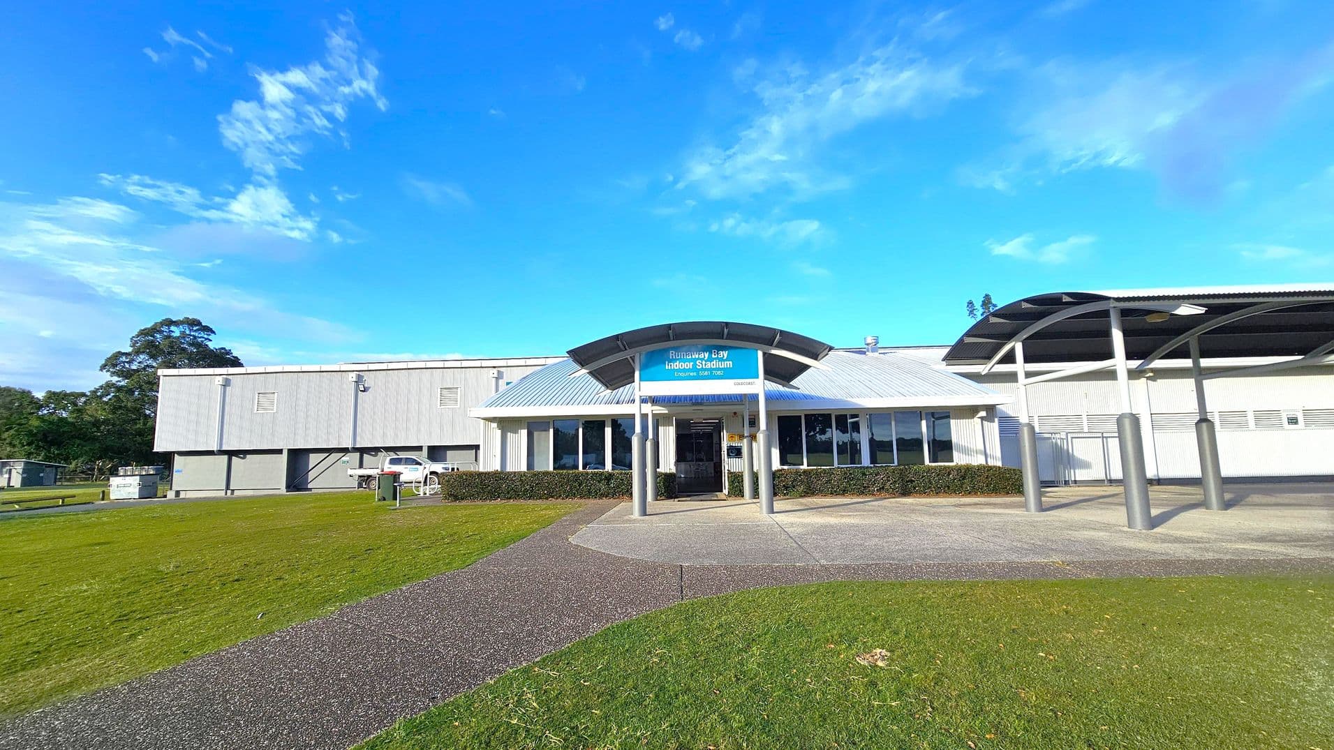 A large building with a metal roof and a sign reading "Runaway Bay Indoor Stadium" above the entrance. The entrance is flanked by glass windows and supported by metal pillars. A covered walkway extends to the right. The foreground features a grassy area with a paved path leading to the entrance. On the left, there is a parked white vehicle and some trees in the background. The sky is clear and blue.