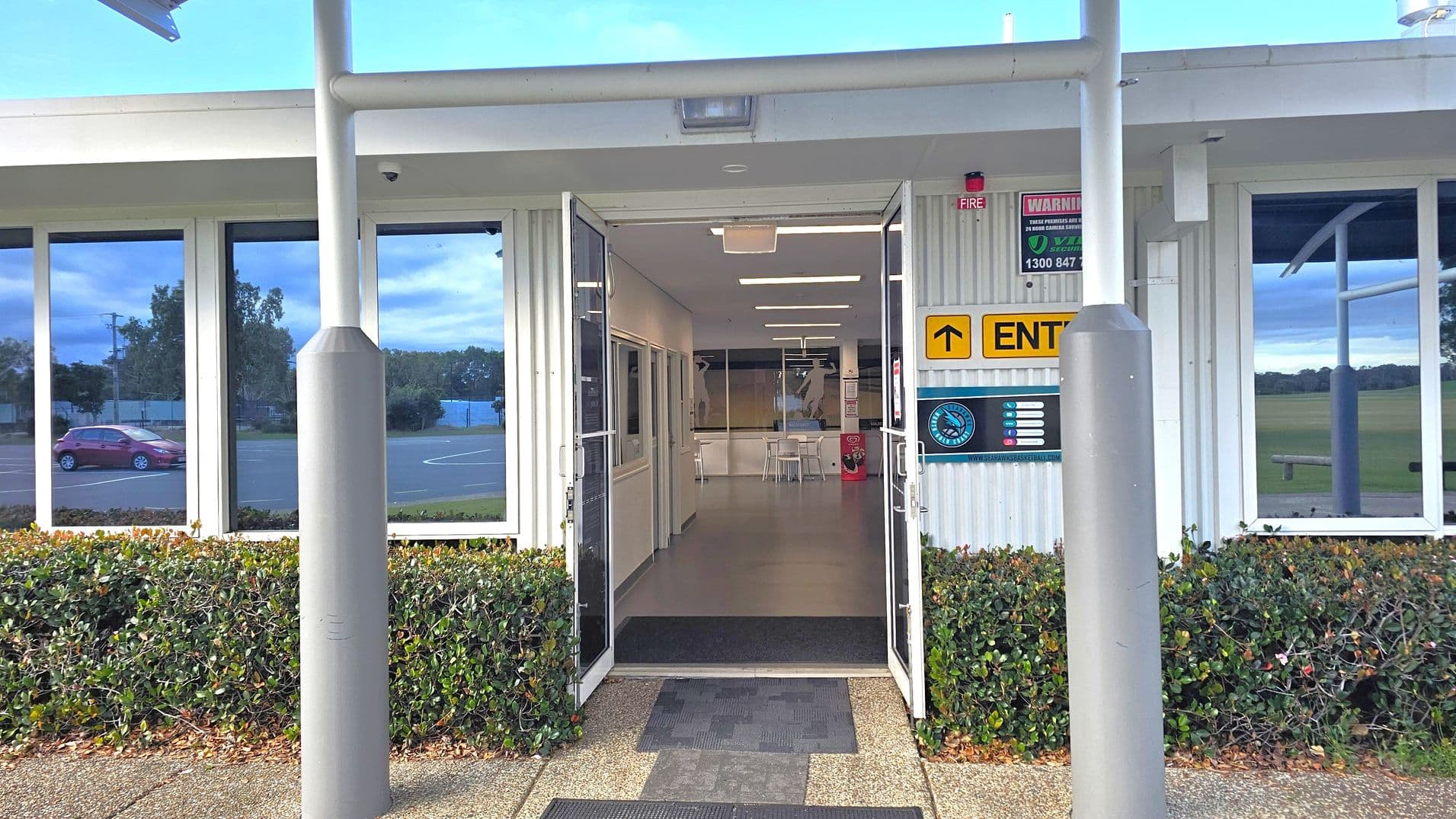 A building entrance with double glass doors open, leading to a hallway with white walls and a grey floor. Above the doors, a yellow sign with an arrow and the word "ENTER" is visible. On the right, a sign with a logo is displayed. The exterior features large windows reflecting a red car and greenery. Two grey bollards stand in front of the entrance, with a hedge lining the path. The ceiling inside has fluorescent lights.