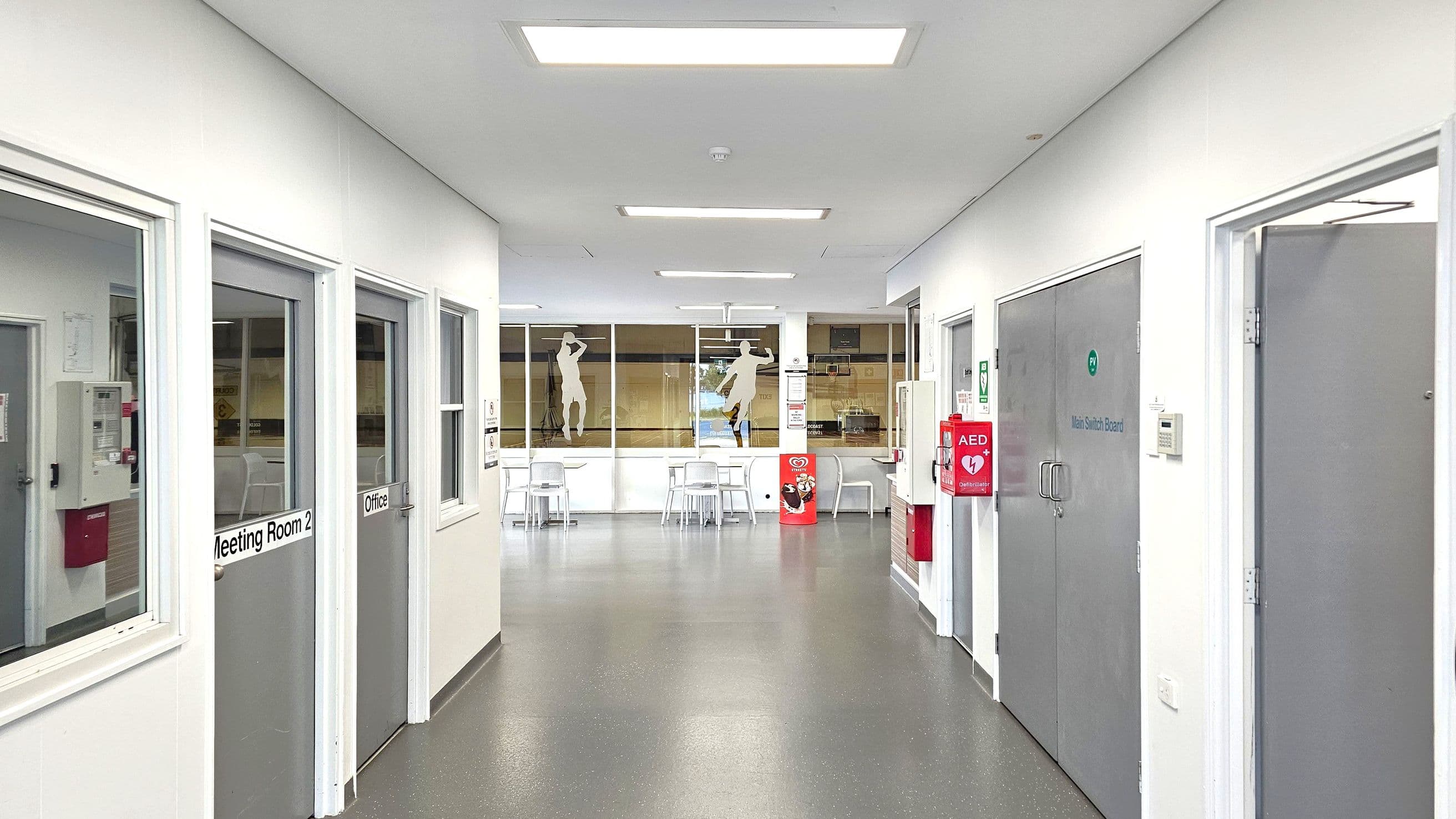 A corridor with grey flooring and white walls. On the left, there are doors labelled "Meeting Room 2" and "Office". On the right, a large grey door is marked "Main Switch Board". An AED device is mounted on the wall. In the background, glass windows display silhouettes of athletes, with tables and chairs visible. The ceiling has rectangular lights.