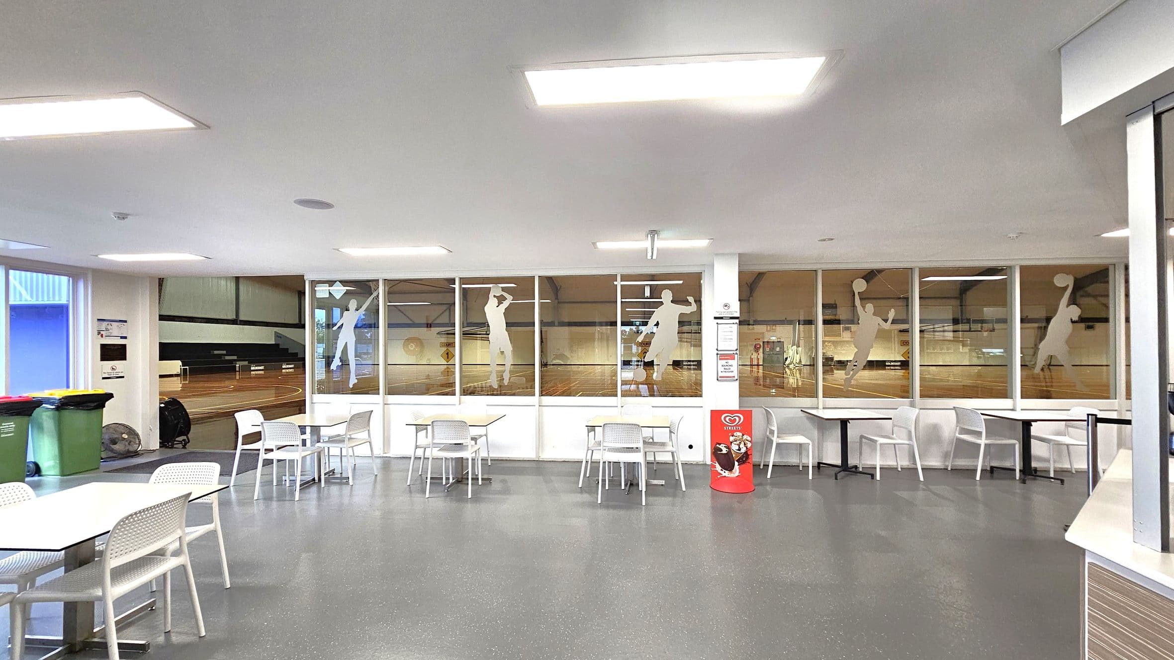 A spacious indoor area with a grey speckled floor and white ceiling featuring rectangular lights. On the left, there are green and yellow bins near a window. In the centre, several white tables and chairs are arranged, with a red ice cream display. The background has large windows with silhouettes of athletes, revealing a wooden sports court with seating.