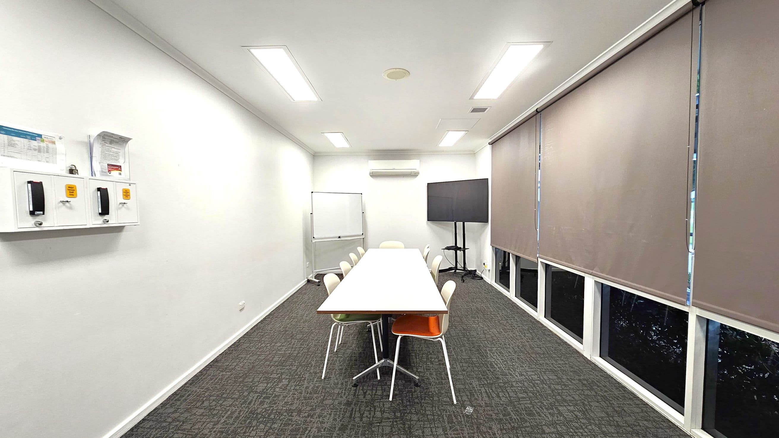 A conference room with a long rectangular table in the centre, surrounded by chairs. On the left wall, there are two wall-mounted boxes with documents and notices. The right side features large windows covered with beige blinds. At the far end, a whiteboard and a large flat-screen TV on a stand are visible. The floor is carpeted in a dark pattern, and the ceiling has recessed lighting and an air conditioning unit.