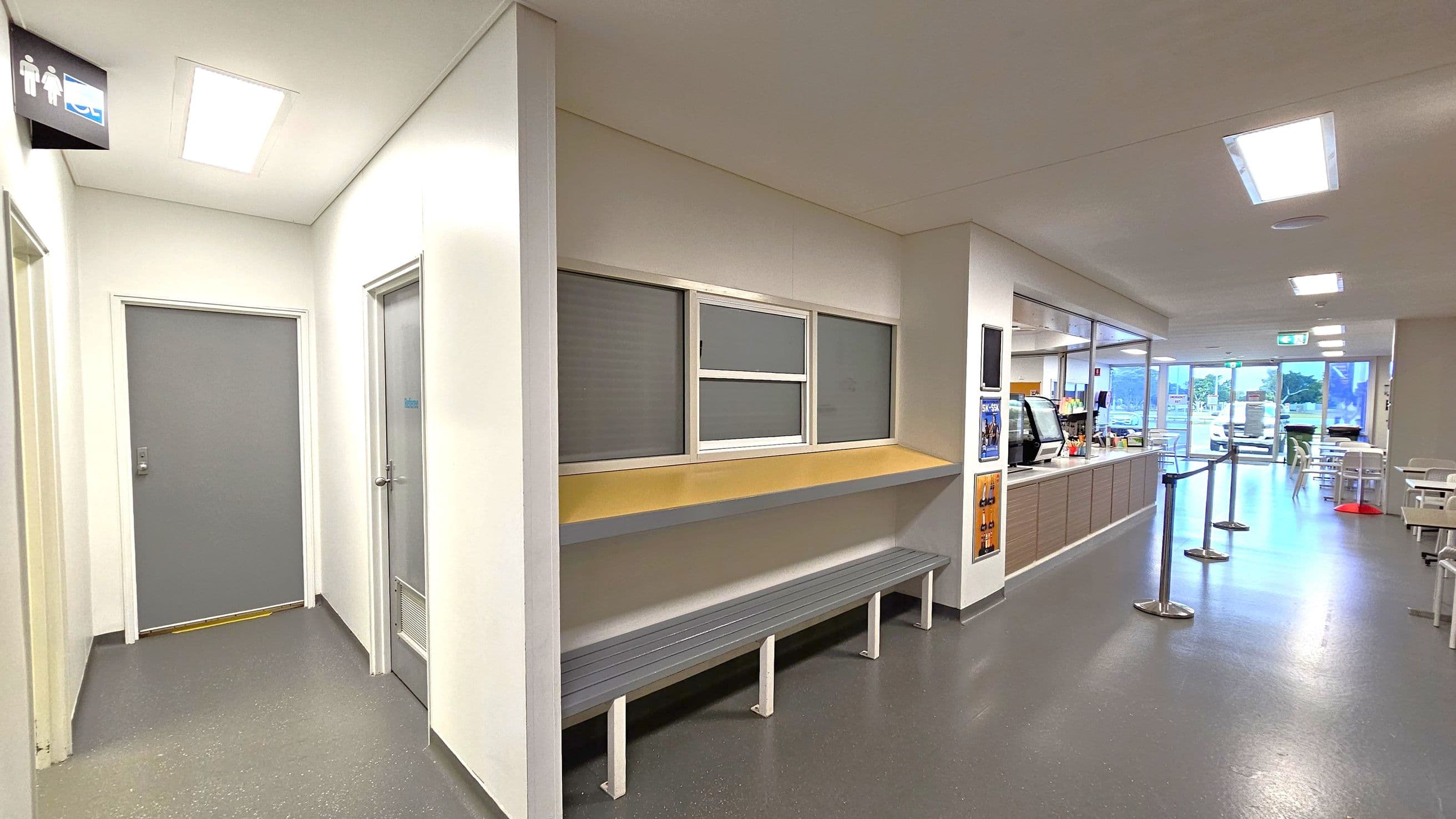A hallway with grey flooring and white walls. On the left, there are two grey doors, one with a sign above indicating toilets. In the middle, a bench is positioned beneath a counter with three windows. On the right, a cafeteria area with a counter, beverage machines, and a queue barrier. Further back, there are tables and chairs near large glass doors leading outside. The ceiling has recessed lighting.
