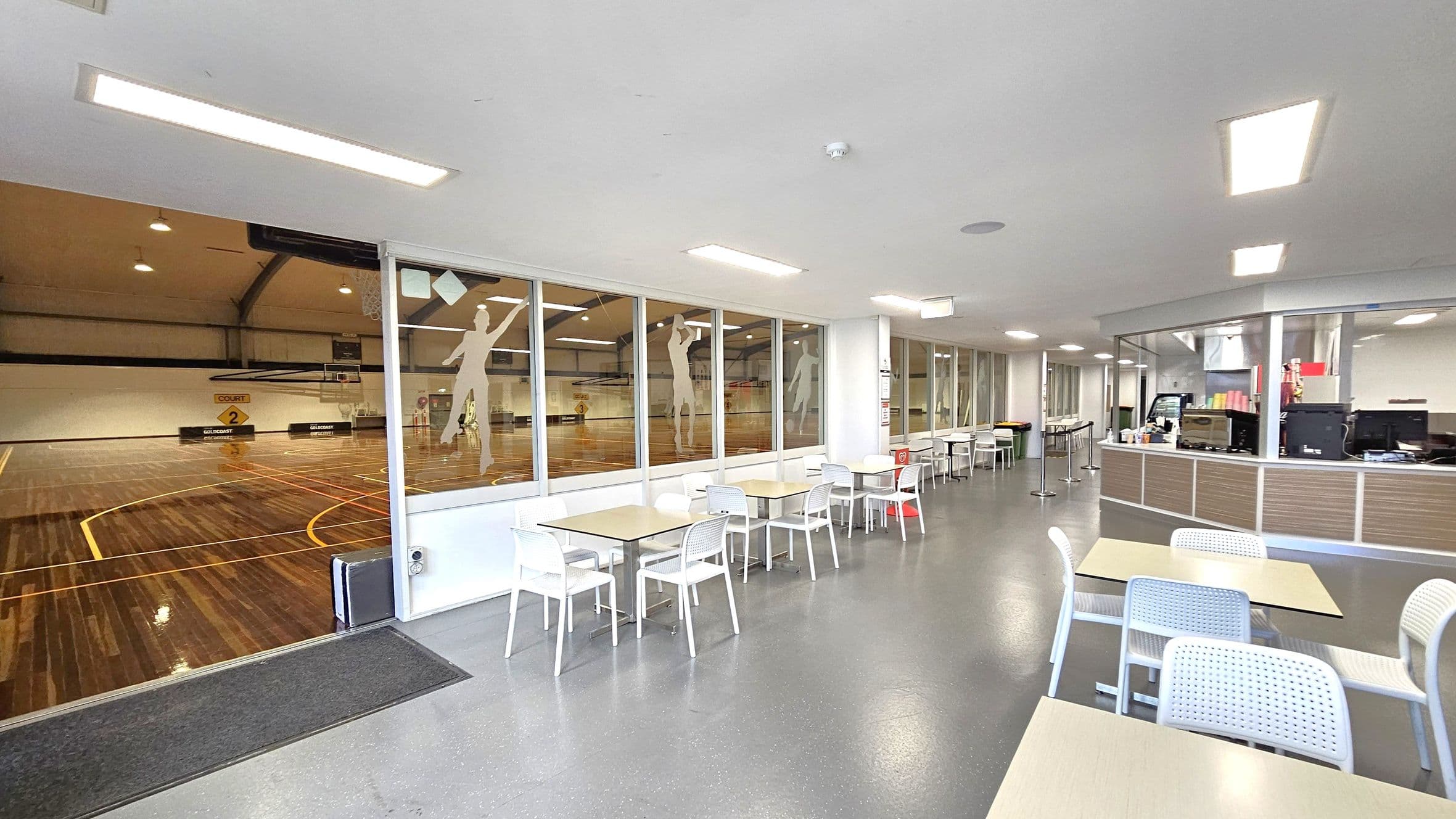 On the left, a large indoor basketball court with polished wooden flooring and yellow lines is visible through glass panels. The middle features a seating area with white tables and chairs on a grey floor. On the right, a counter with computers and various items is present, possibly a reception or café area. The ceiling is white with rectangular lights.