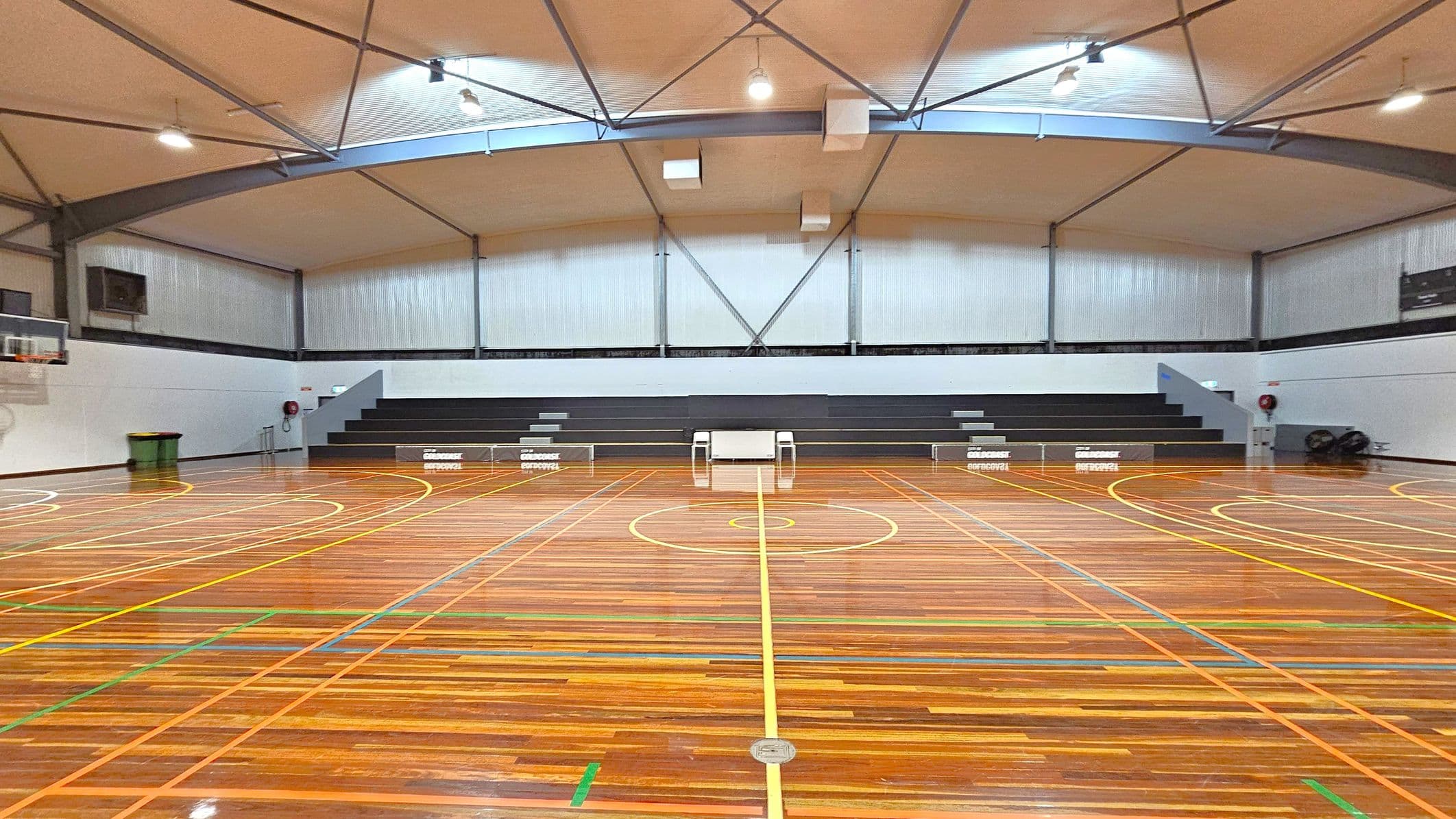 A spacious indoor sports court with a polished wooden floor marked with yellow, green, and blue lines. In the background, there are grey bleachers with a small table and chairs in the centre. The walls are light grey with corrugated metal panels. The ceiling is high with exposed beams and several lights. On the left, a basketball hoop is visible, and a green bin is placed nearby. On the right, a fire extinguisher is mounted on the wall.