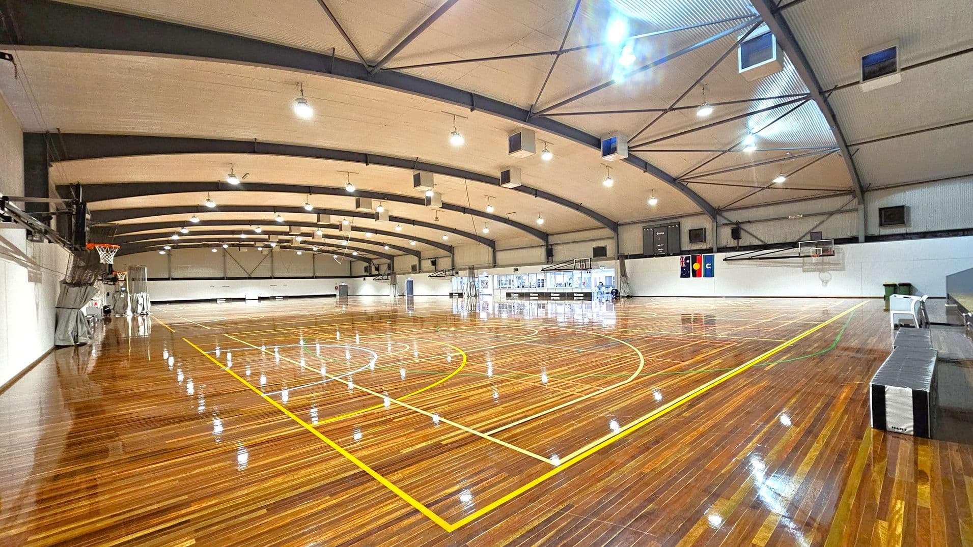 A spacious indoor basketball court with a polished wooden floor marked with yellow lines. On the left, a basketball hoop is visible, and on the right, there are benches and a scoreboard. The ceiling is high with exposed beams and multiple lights. The walls are white, and there are flags displayed on the far wall. The court is well-lit, and the space is open and empty.