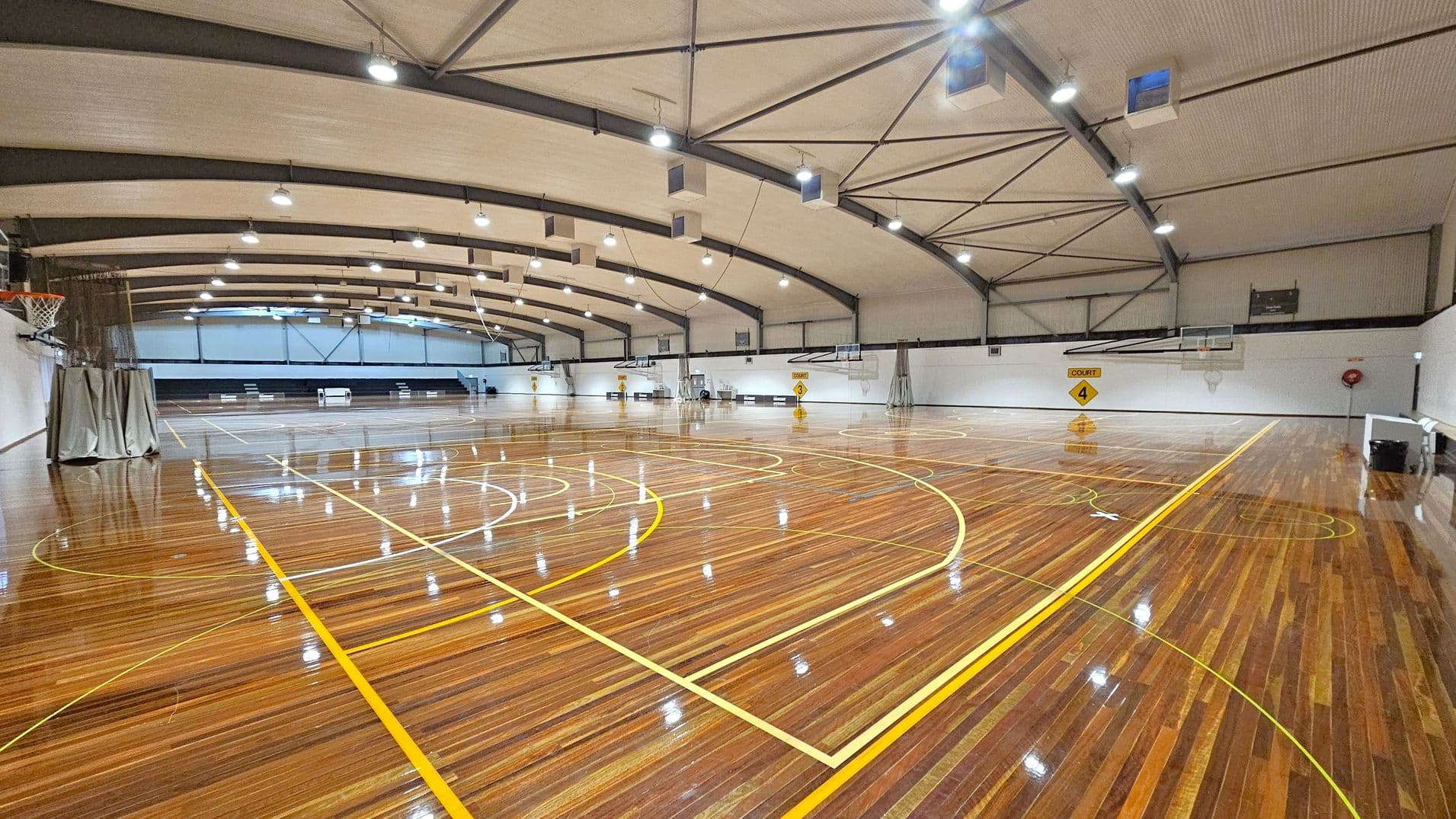 A spacious indoor basketball court with a polished wooden floor marked with yellow lines. On the left, a basketball hoop is visible with a net and a grey curtain. The ceiling is arched with exposed beams and multiple lights. The right side has another basketball hoop and a sign indicating "Court 4". The background features a white wall with additional hoops and a small set of bleachers. The area is well-lit, and the floor reflects the overhead lights.