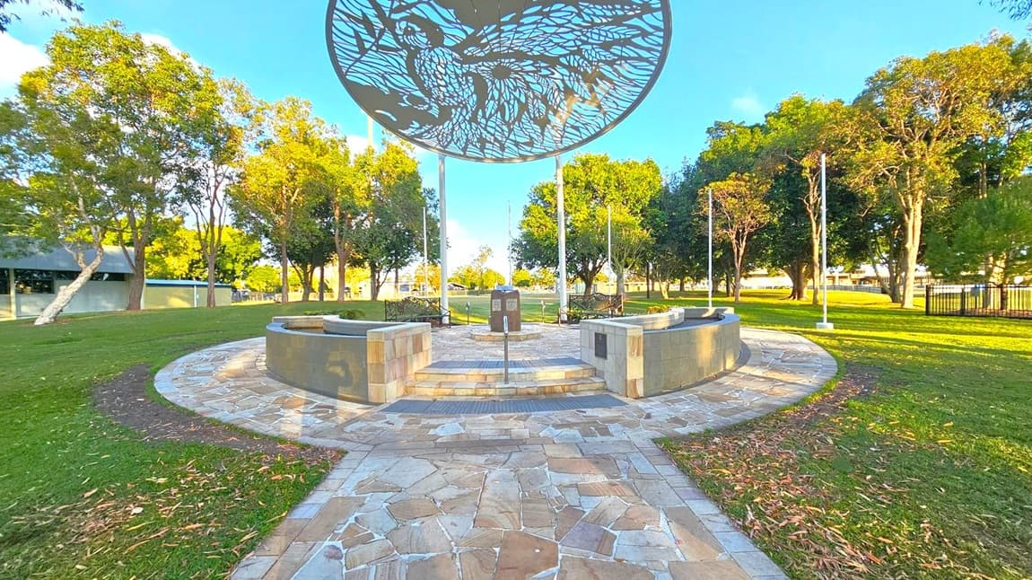 A circular stone pathway leads to a central monument with a decorative metal canopy above, featuring intricate cut-out designs. The area is surrounded by a low stone wall and benches. Trees with lush green foliage line the background, and a building with a sloped roof is visible to the left. The ground is covered with grass, and there are scattered leaves on the path.