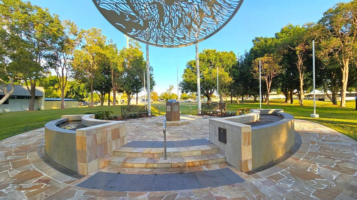 A circular paved area with a central monument surrounded by curved stone benches. Above, a large metal canopy with intricate cut-out patterns is supported by poles. The foreground features stone steps leading up to the monument, with a metal handrail. Benches and flagpoles are positioned around the circle. The background is a grassy park with numerous trees and a few buildings visible in the distance.