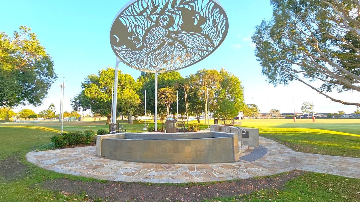 A circular stone platform with a decorative metal canopy featuring intricate patterns is in the centre. The platform is surrounded by a paved walkway. Trees are scattered around the area, with a grassy field extending into the background. Rugby posts are visible in the distance. The sky is clear, and the scene is well-lit, suggesting daytime.