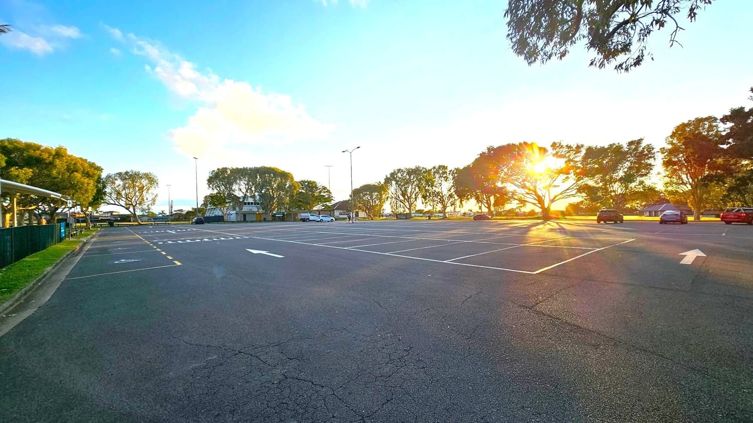 An empty car park with marked spaces and a few parked cars on the right. The sun is setting behind a row of trees, casting long shadows across the asphalt. On the left, a green fence runs alongside a grassy area. In the background, there are more trees and a few buildings. The sky is clear with some clouds, and the overall scene is calm and quiet.
