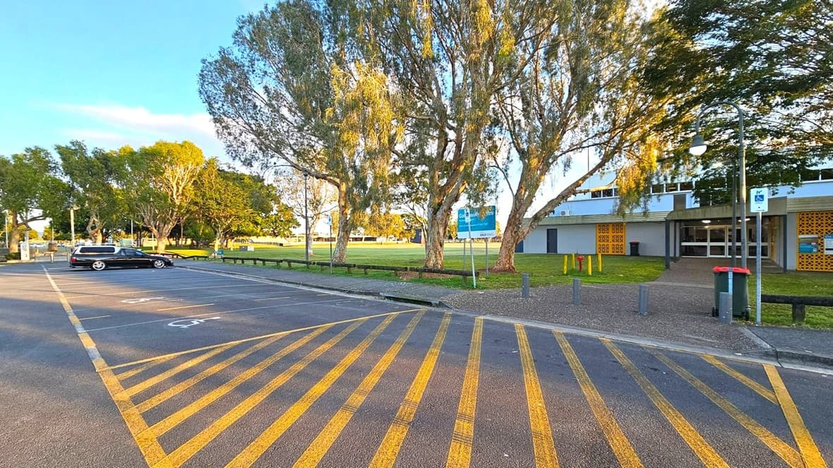 A car park with yellow diagonal lines on the ground is in the foreground. A single black car is parked on the left. In the middle, there are several large trees with green foliage. On the right, a building with a grey facade and yellow decorative panels is visible. A pathway leads to the entrance, and a green bin is nearby. A signpost and a lamp post are also present. The background features more trees and an open grassy area.