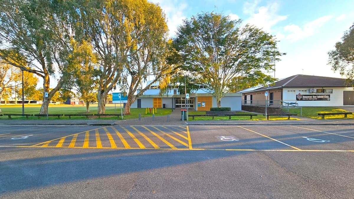 A paved parking area with yellow-striped markings and designated disabled parking spaces in the foreground. In the middle, a building with a sign reading "Runaway Bay Soccer Club" is partially visible on the right. The building is surrounded by trees, with a grassy area and a blue sign on the left. Benches and a pathway lead to the entrance of the building. The scene is well-lit, suggesting daylight.