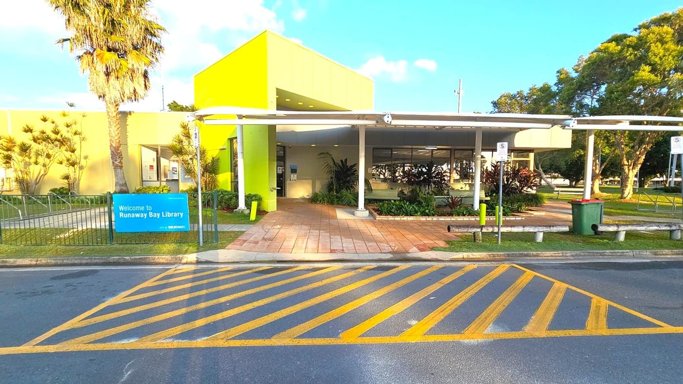 A library entrance with a bright green facade and a flat roof. A paved walkway leads to the entrance, flanked by plants and a small garden. A blue sign on the left reads "Welcome to Runaway Bay Library." A palm tree and other greenery are visible on the left. The foreground features a road with yellow diagonal lines. On the right, there are parking signs and a green bin. Trees are visible in the background.