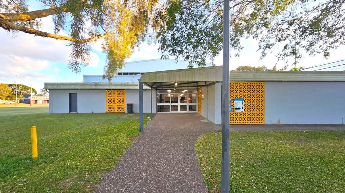 A building with a flat roof and a covered entrance in the centre. The entrance has glass doors and is flanked by yellow lattice panels on both sides. A pathway leads up to the entrance, bordered by grass. A tree branch extends from the left, casting shadows on the building. A yellow bollard is visible on the left side of the grass. The building has a grey exterior with a small dark door on the left.