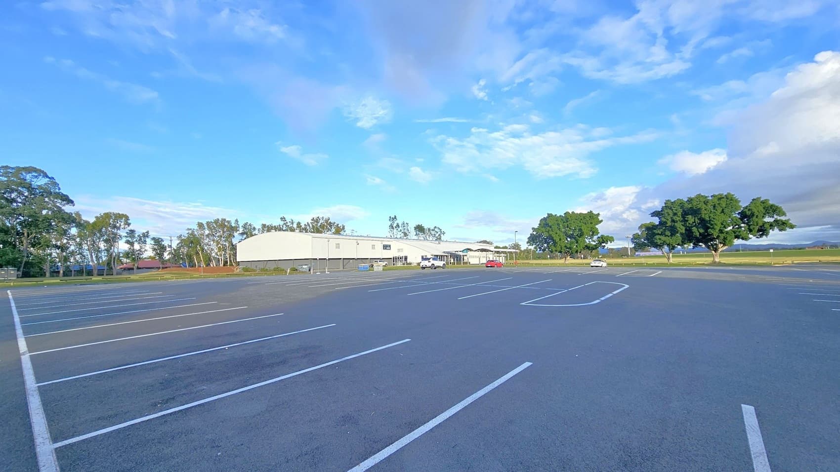 An expansive, empty car park with white lines marking parking spaces. In the background, a large, light-coloured building is visible, surrounded by trees. A few vehicles are parked near the building. To the right, there are large, leafy trees and open fields. The sky is mostly clear with some clouds.