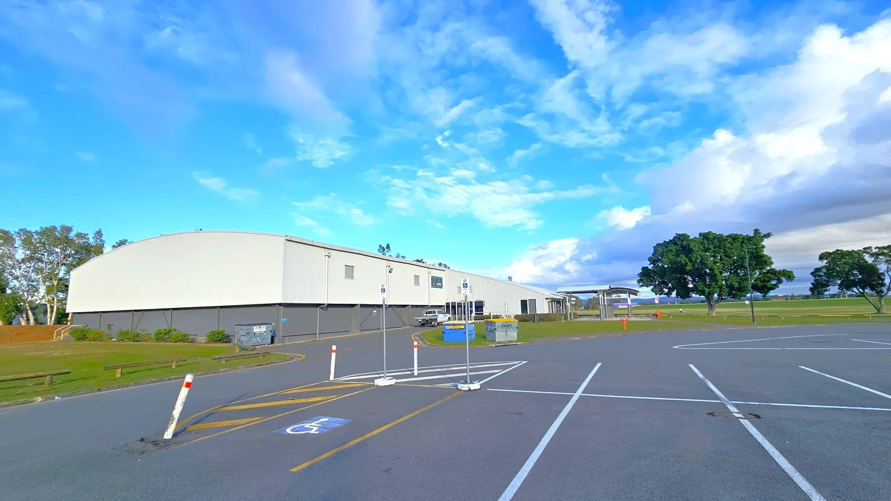 A large, white industrial building is on the left, with a grey lower section and a few small windows. In front of it, a car is parked near a blue dumpster. The foreground features a car park with marked spaces, including a disabled parking spot. On the right, there are open grassy fields with a few large trees. The sky is mostly clear with some clouds.