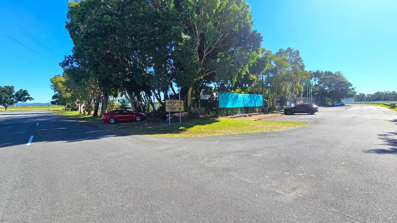 A paved parking area with a few cars is on the right. On the left, a road runs alongside a row of large trees providing shade. A signpost near the trees indicates directions for cricket and tennis. A blue banner is partially visible among the trees. The background features open grassy fields and more trees, with a clear blue sky above.