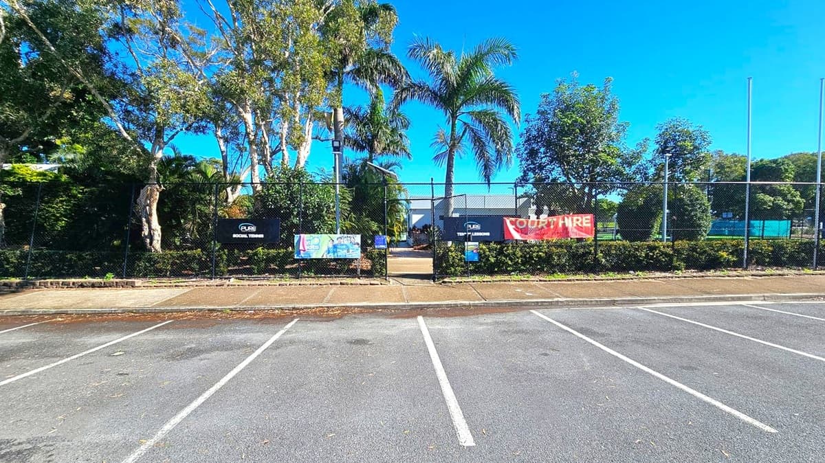 A tennis court entrance is visible, surrounded by a black chain-link fence with several signs and banners attached. The foreground features an empty parking area with marked spaces. Behind the fence, there are tall trees, including palm trees, and dense greenery. The pathway leading to the entrance is flanked by hedges. The sky is clear and blue.